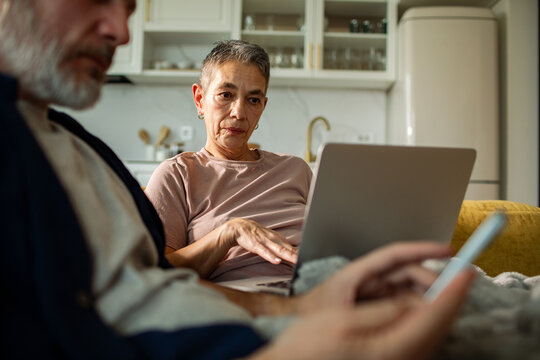 Senior woman using laptop at home while partner checks phone in kitchen
