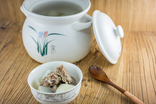 A serving of pork rib and radish soup in a bowl, with a clay pot and lid on a wooden table.