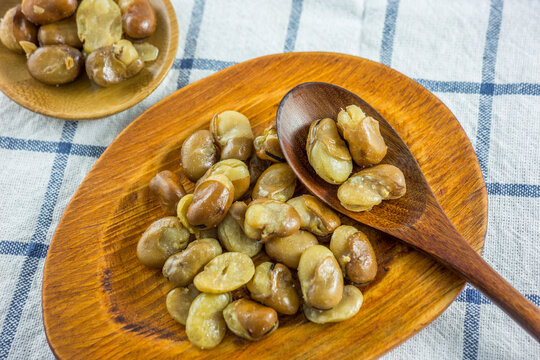 Roasted cooked broad beans presented on wooden plates and a spoon, placed on a checkered tablecloth.