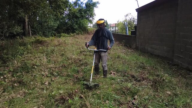 Professional gardener wearing protective gear and using a powerful string trimmer to cut down overgrown grass and weeds in a backyard, performing seasonal lawn care and maintenance. 
