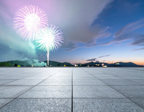 Firework display above empty tiled plaza at blue hour with glowing bursts, soft smoke, distant mountain silhouette, and colorful town lights creating spacious festive scene