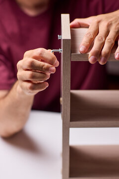Young man assembling wooden shelf in indoor workspace, close-up view of hands tightening hardware with drill, warm lighting and focused expression during DIY repair task