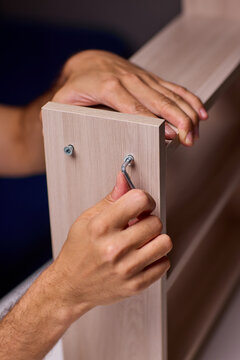 Young man assembling wooden shelf in indoor workspace, close-up view of hands tightening hardware with drill, warm lighting and focused expression during DIY repair task