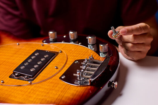 Right hand placing guitar string tuning peg on electric guitar bridge with amber sunburst finish, chrome hardware, glossy wood grain, and warm closeup light