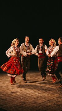 Ensemble of dancers in traditional Bulgarian folk costumes performing a lively dance on a wooden stage