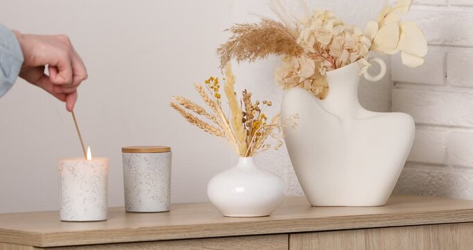 Woman lighting candle on wooden dresser near vases of dry flowers indoors, closeup
