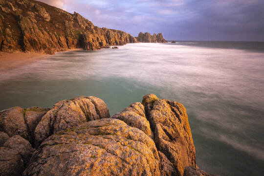 View of Porthcurno Beach with rugged cliffs, golden hour light on rocks, and long exposure sea in Porthcurno, England, United Kingdom.