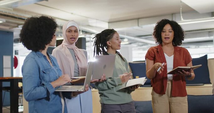 Diverse team open stand-up, woman rust top with notes, speaking, team noting laptops