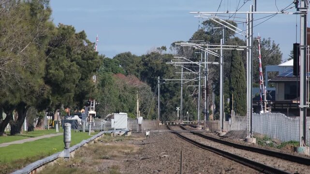 Railway tracks leading toward a level crossing with safety boom gates, overhead power lines, and residential houses in Seaholme, Melbourne, Victoria, Australia. Suburban rail infrastructure 