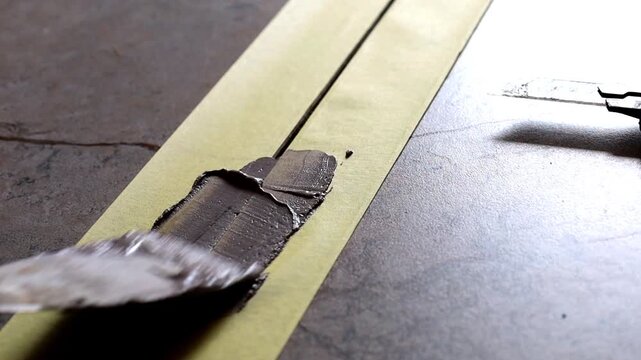 Professional craftsman carefully applies putty or special adhesive using a spatula along protective masking tape for precise surface repair during construction and finishing works indoors.