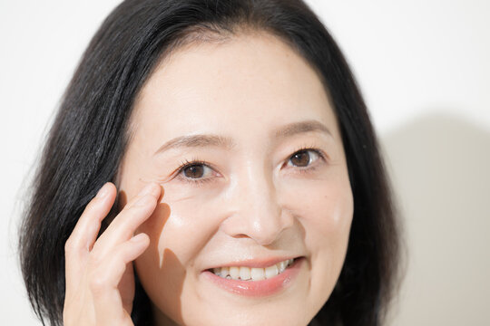 目元のシワをチェックしながら笑顔を見せる日本人ミドル女性、50代のエイジングケアとスキンケアイメージクローズアップ