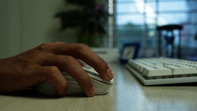 Hand Using Computer Mouse at Office Desk, Close-up of a hand using a computer mouse beside a keyboard on a desk, with soft window light and a blurred office background.