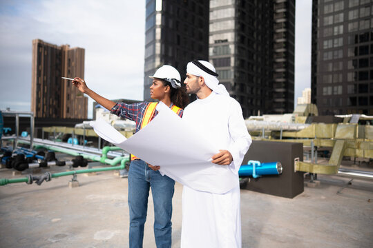 Engineer and client reading the plant drawings on site as the large blueprint is unrolled open between them, with insulated pipework and blue valves filling the roof behind.