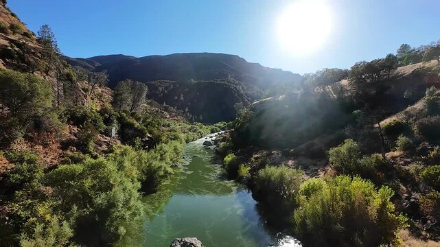 Cinematic first-person view of a drone flying forward along the beautiful Cache Creek in California, navigating through a lush, green canyon with the bright sun shining in the sky
