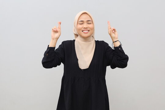 A young Asian woman wearing a headscarf smiles with her eyes closed and fingers crossed, making a wish, posing against a plain white background.