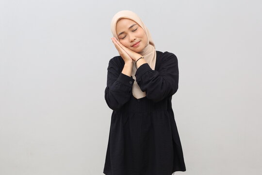 A young Asian woman wearing a headscarf stands against a white background, miming a sleeping gesture with an adorable expression, smiling and closing her eyes.