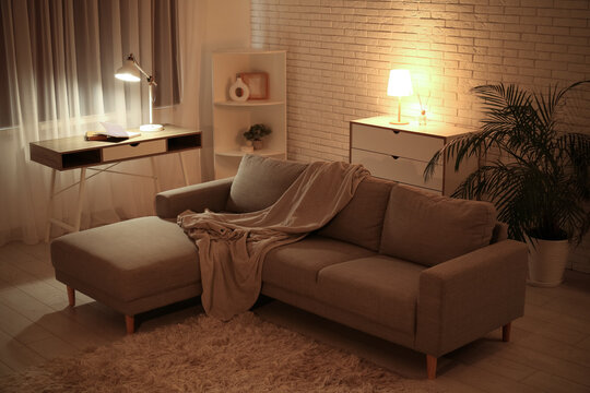 Interior of night living room with sofa, shelving unit, glowing lamps, commode and workplace