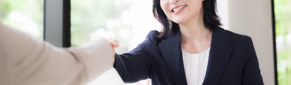 笑顔で握手をする日本人ミドル女性、商談や契約で相手と信頼関係を築く50代女性ビジネスパーソン顔無しのバナー
