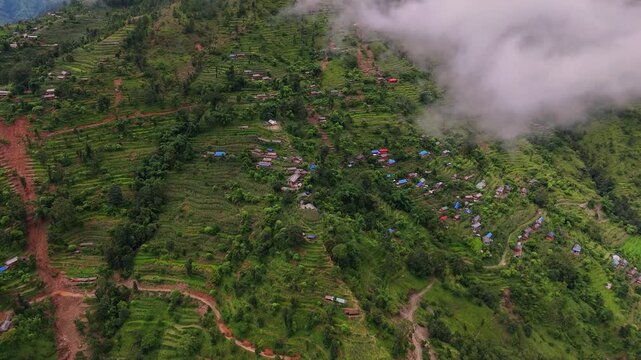 Aerial view of a steep green terraced hillside dotted with rural homes and a winding dirt road under low mountain fog in Nepal