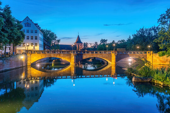 Stunning evening blue hour view of the illuminated historic stone arch bridge spanning the calm Pegnitz River with glowing architectural reflections in Nuremberg
