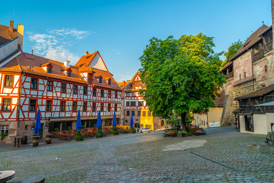 square featuring traditional half-timbered medieval buildings, outdoor dining tables, and a large green tree in the old town of Nuremberg
