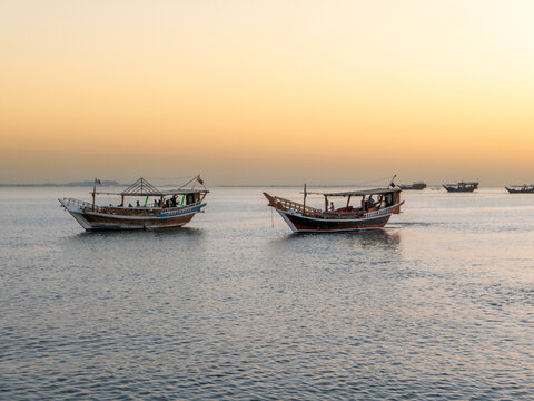 Traditional wooden dhow boats floating on calm waters in Doha Bay with warm golden sunset sky in background.