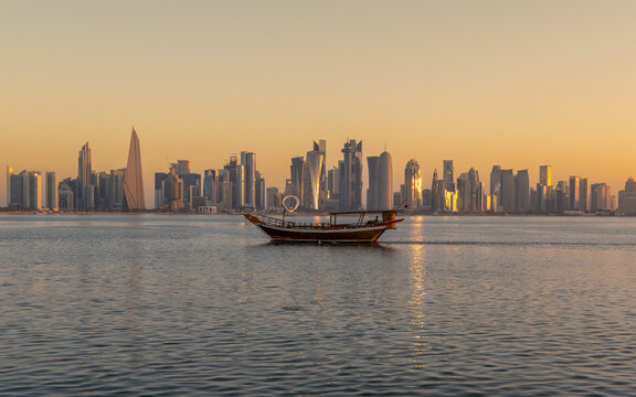 Modern West Bay skyline in Doha featuring highrise skyscrapers and traditional dhow boat on calm waters at golden sunset.
