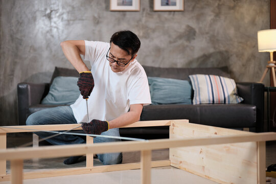 Asian mature man concentrates on self-assembling wooden furniture with DIY tools, tightening strength with a screwdriver, home improvement, hobby carpentry workshop, expert handyman, active lifestyle.