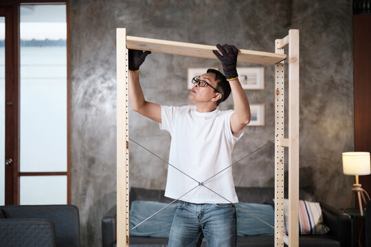 Asian mature man in gloves determined to assemble furniture with DIY tools, installing strong wooden shelf, home improvement, craft making, hobby carpentry workshop, expert handyman, active lifestyle.