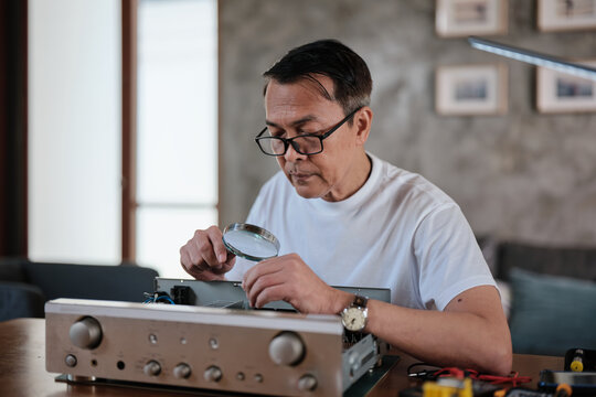 Mature Asian man disassembling and checking audio equipment, looking with magnifying glass at circuit board details for repair, home appliance DIY fix, electronics component hobby, handyman workshop.