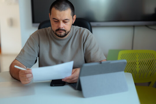 Concentrated Male Teacher Reading Paper Document In Front Of Tablet Screen. Focused man sits at his desk analyzing documents during online class