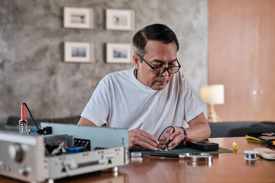 Mature Asian man voltage test at disassembling circuit board with multimeter tool, focusing on precision checks for home electrical appliance DIY fix, electronics component hobby, handyman workshop.