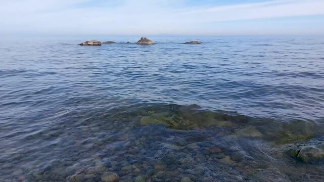 Clear lake water with visible pebbles on the bottom and rocks in the distance under a blue sky, likely Lake Baikal