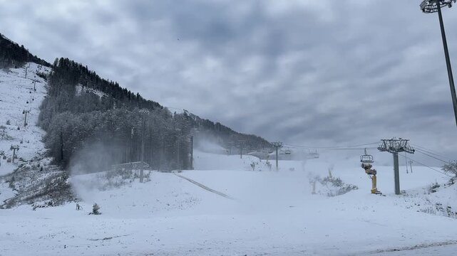 Snow-covered ski resort with empty slopes and ski lifts. Cloudy sky and frosty trees create a winter landscape.