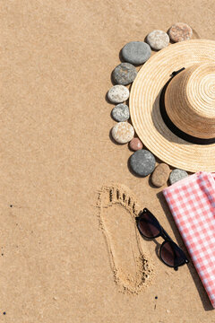 Beach straw hat resting on golden sand surrounded by smooth round pebbles alongside sunglasses and checkered towel arranged overhead for vacation concept overhead flat lay composition view