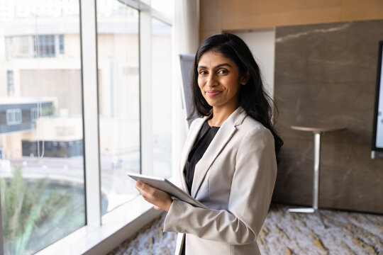Confident Indian businesswoman standing in modern office holding digital tablet, looking at camera. Corporate professional using technology for business management, data review and digital workflow
