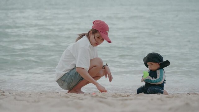 Happy mother and adorable baby boy sitting on sandy beach together during summer vacation