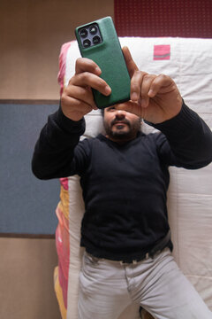 A top-down, vertical view of a cell phone held in the hands of a man who is lying on his bed in his bedroom while taking a moment of leisure and relaxation at home