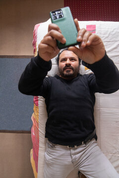A man lying on a bed in a bedroom with his arms outstretched, holding and looking at his cell phone screen