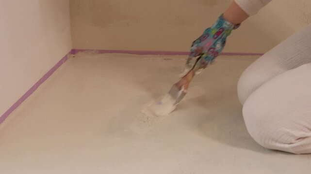 A worker kneels on the floor, dips a brush into a paint tray, and applies primer along the edge near a wall marked with tape.