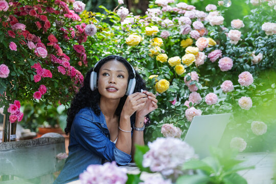 A woman wearing headphones sits among colorful blooming roses and lush greenery, looking upward in a sunlit garden. The peaceful scene captures personal downtime, outdoor relaxation