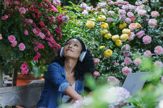 A woman wearing headphones sits among colorful blooming roses and lush greenery, looking upward in a sunlit garden. The peaceful scene captures personal downtime, outdoor relaxation