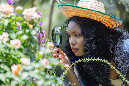 A woman wearing a straw hat closely examines garden flowers with a magnifying glass. Her focused expression highlights attention to detail, thoughtful plant care, and curiosity about nature.