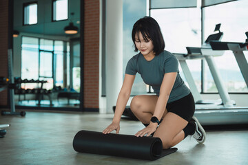 Fototapeta premium Young woman rolling fitness mat on gym floor preparing for workout session in modern exercise room with equipment