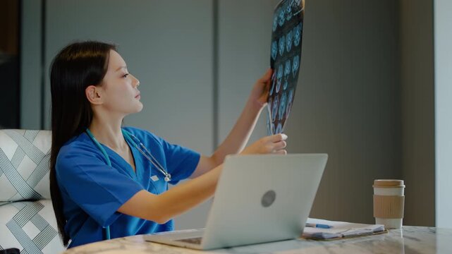 Young Asian female doctor analyzing brain x-ray CT scan film while typing on laptop at desk in hospital office. Professional medical worker reviewing patient radiograph and health data online.
