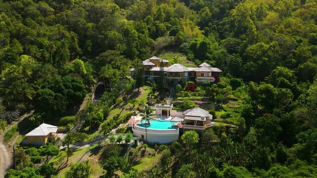 Slow left pan across Beau Estate Domain in Saint Lucia, a private luxury villa with infinity pool, terraced gardens and pyramid roofs set into dense green Caribbean rainforest hillside