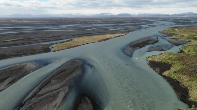 Braided glacial channels divide dark glaciofluvial sediment across Skeidararsandur, with vegetated river margins and extensive outwash plain stretching toward distant mountains, Southeast Iceland, dro