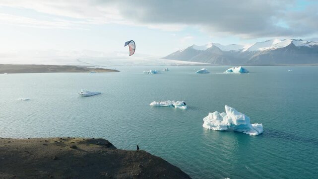 Kite surfer flies above iceberg filled J&ouml;kuls&aacute;rl&oacute;n Glacier Lagoon, with dark volcanic shoreline and broad mountain ridge surrounding glacial waters in Southeast Iceland, drone aerial shot.