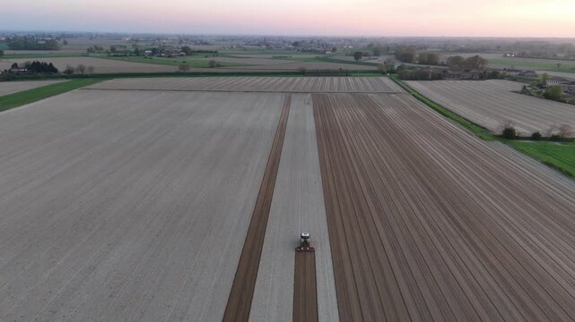 Tractor performs precise parallel field passes across cultivated farmland, creating uniform soil bands within large agricultural parcels across Po Valley, Emilia-Romagna, Italy, aerial view.