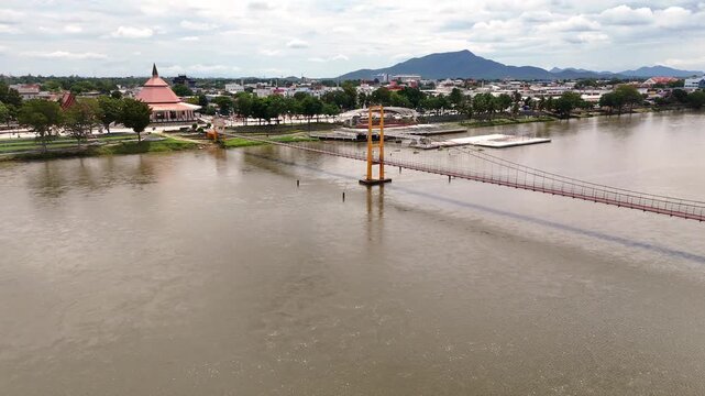 An aerial panning video sequence of the Somphot Krung Rattanakosin 200 Pee suspension bridge, crossing a wide muddy river towards a Thai town with a prominent temple and distant mountains under a clou
